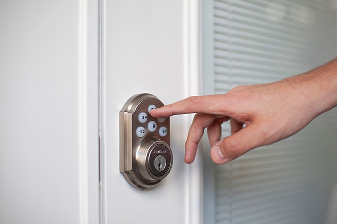 Hand reaching out to push a number on a smart lock installed on a white front door.