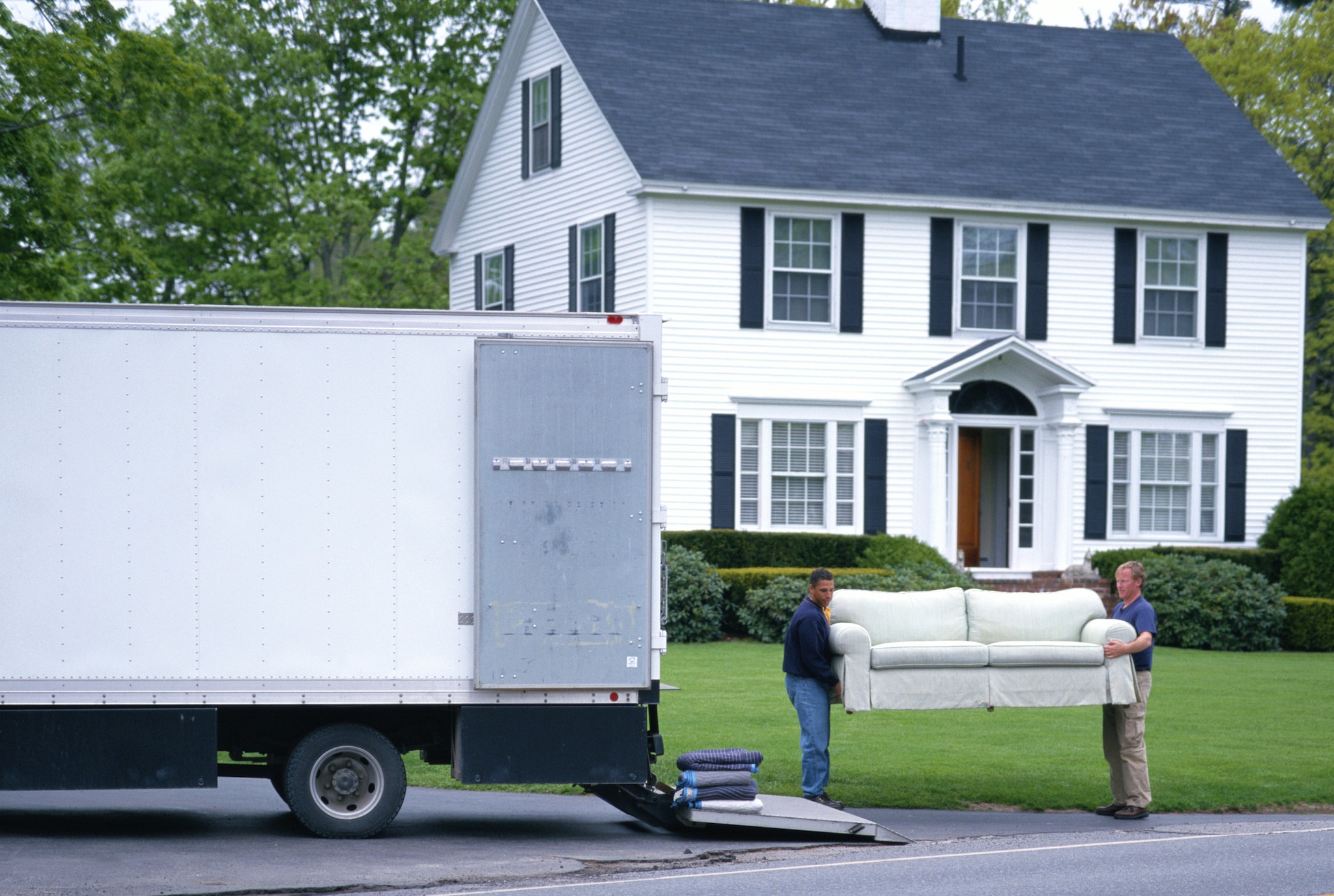 Two men move a white loveseat into a large moving truck in front of a white, two-story home.