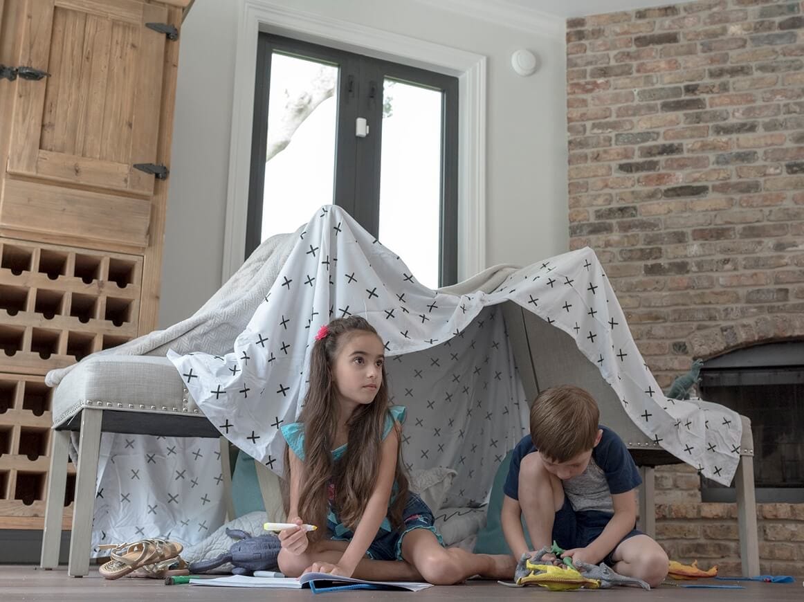 Two children coloring and playing with toys under a sheet draped over two chairs with an ADT smoke detector in the room