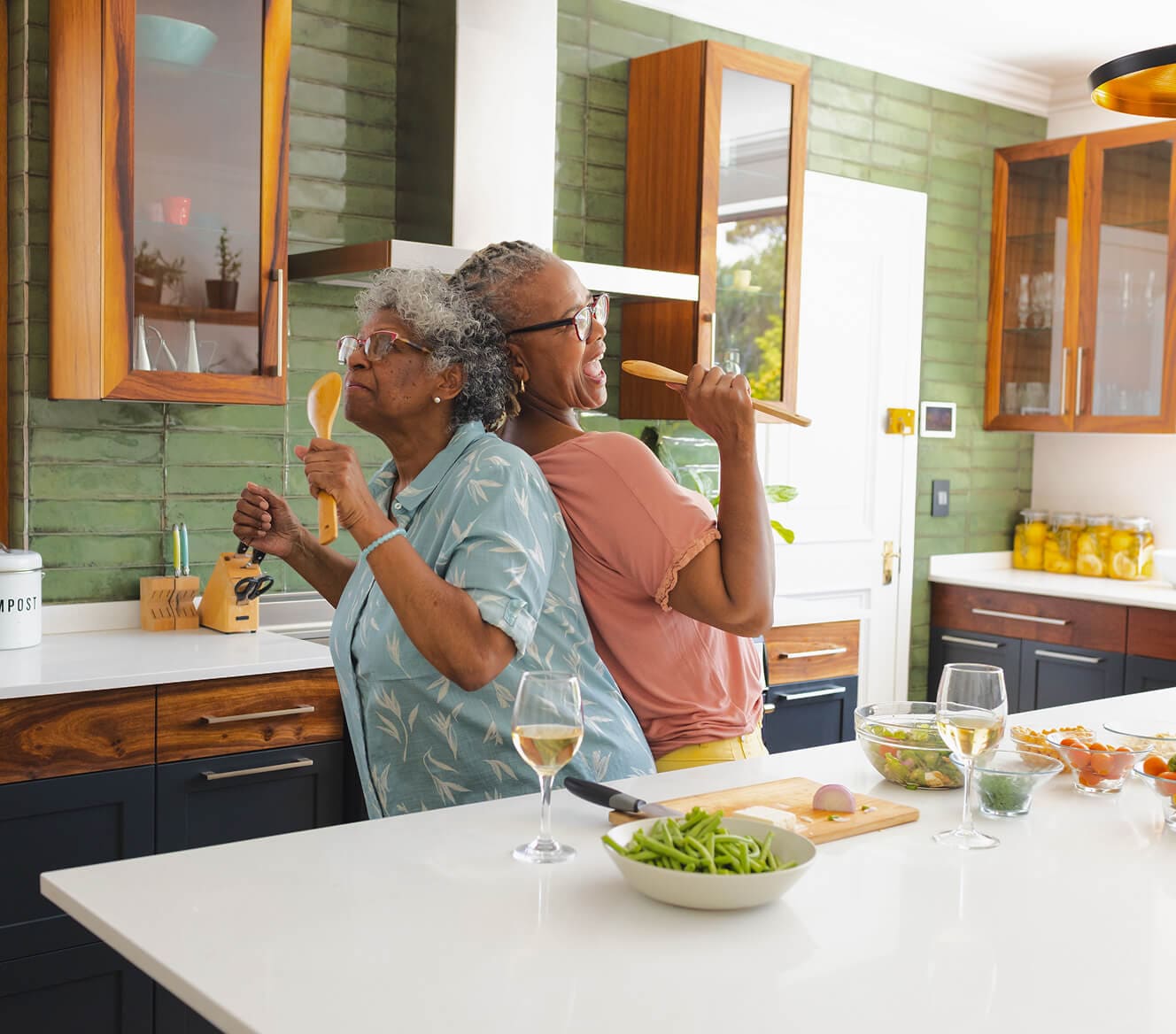 Two joyful older women in a modern kitchen, standing back-to-back while singing into wooden cooking utensils like microphones. They are surrounded by fresh ingredients and glasses of white wine enjoying a fun and lighthearted moment while cooking together