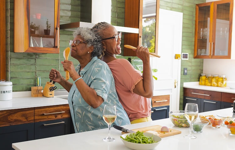 Two joyful older women in a modern kitchen, standing back-to-back while singing into wooden cooking utensils like microphones. They are surrounded by fresh ingredients and glasses of white wine enjoying a fun and lighthearted moment while cooking together