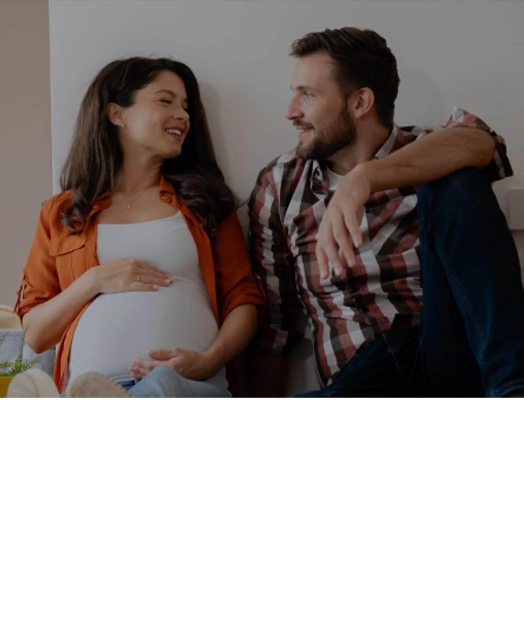 Young couple sitting among moving boxes, smiling and talking, as the woman cradles her pregnant belly—suggesting a new beginning in their new home.