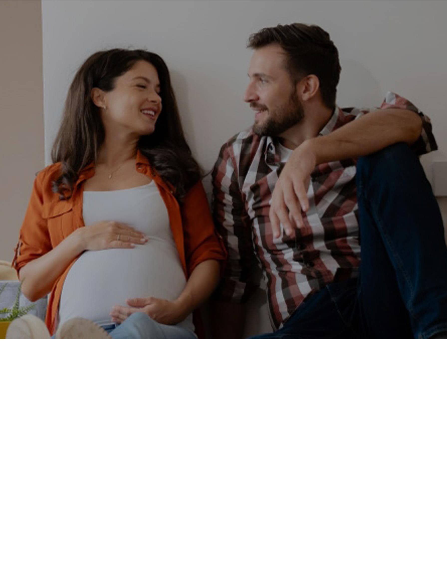 A pregnant woman and a man sitting on the floor of a new apartment, surrounded by cardboard boxes and household items, smiling at each other during the moving process.