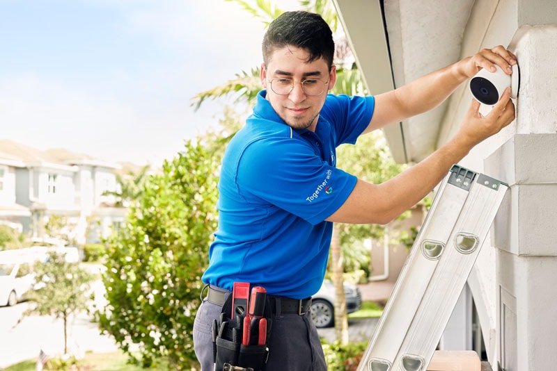 ADT technician in blue uniform installing a smart outdoor security camera on the exterior wall of a home