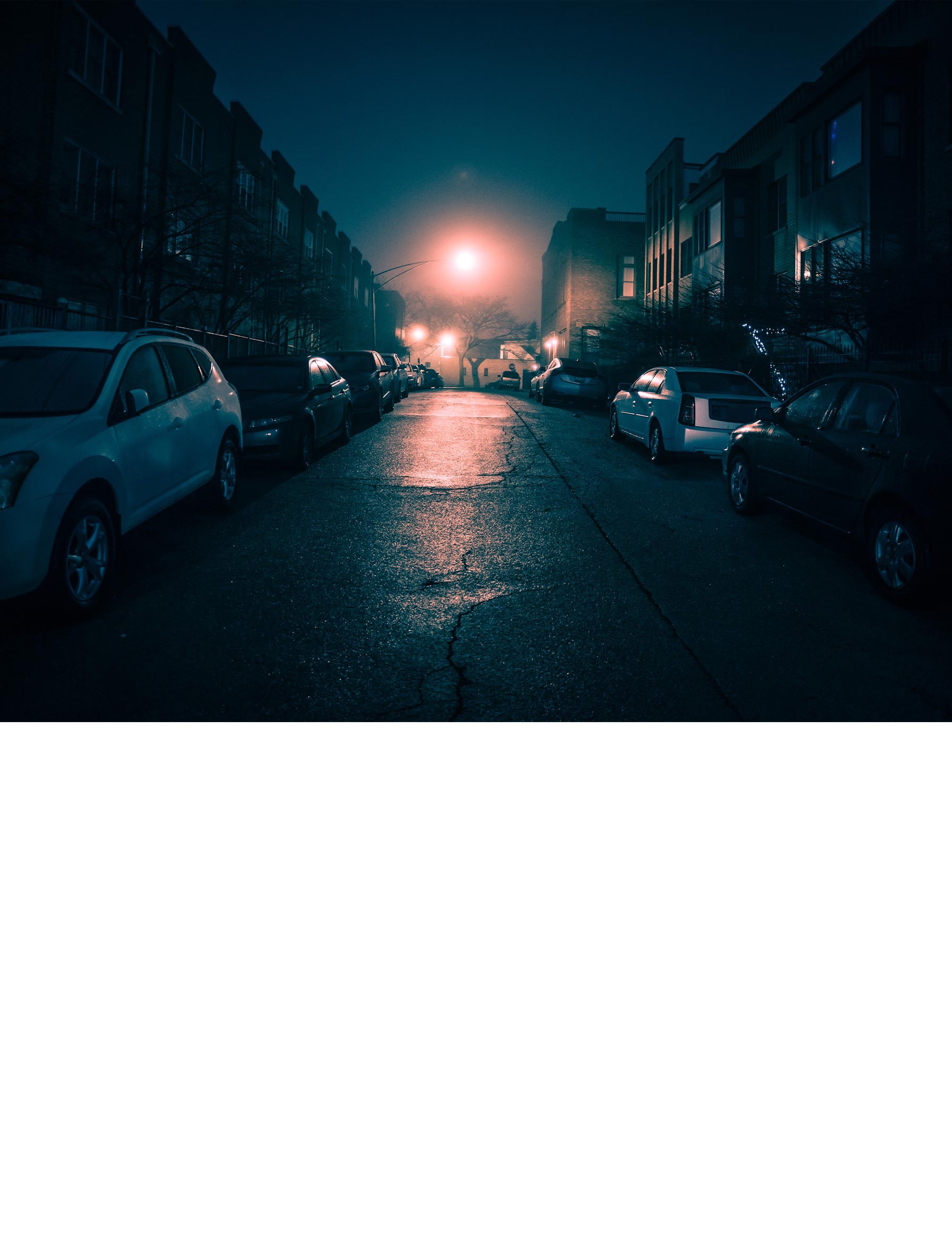 Moody nighttime city street with parked cars, wet pavement, and glowing streetlights in a foggy urban neighborhood.