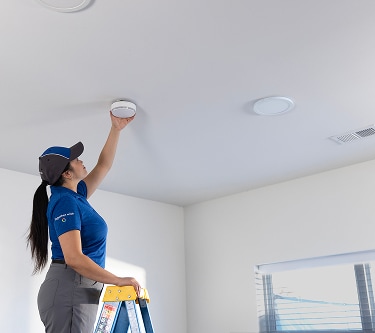 A female technician on a step ladder installs a smoke detector on the ceiling inside a modern home.