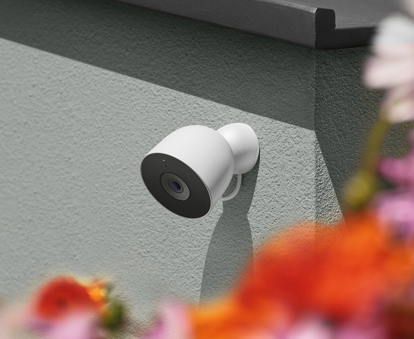 White outdoor security camera mounted on a house wall above blurred red and orange flowers White outdoor security camera mounted on a house wall above blurred red and orange flowers