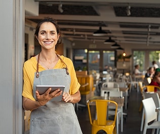 A smiling business owner in a yellow shirt and apron stands in a well-lit café, holding a tablet to manage operations.