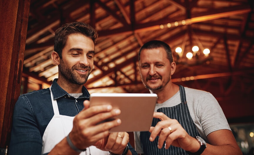 Two men in aprons smile while reviewing lighting information on a tablet, representing small business owners using smart tools.