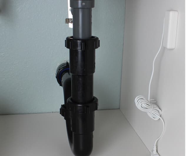 A flood sensor is mounted under a sink with visible plumbing, detecting leaks to help prevent damage.