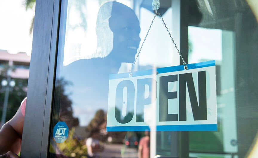 A hanging “Open” sign on a glass door with an ADT security sticker, and the reflection of a smiling customer.
