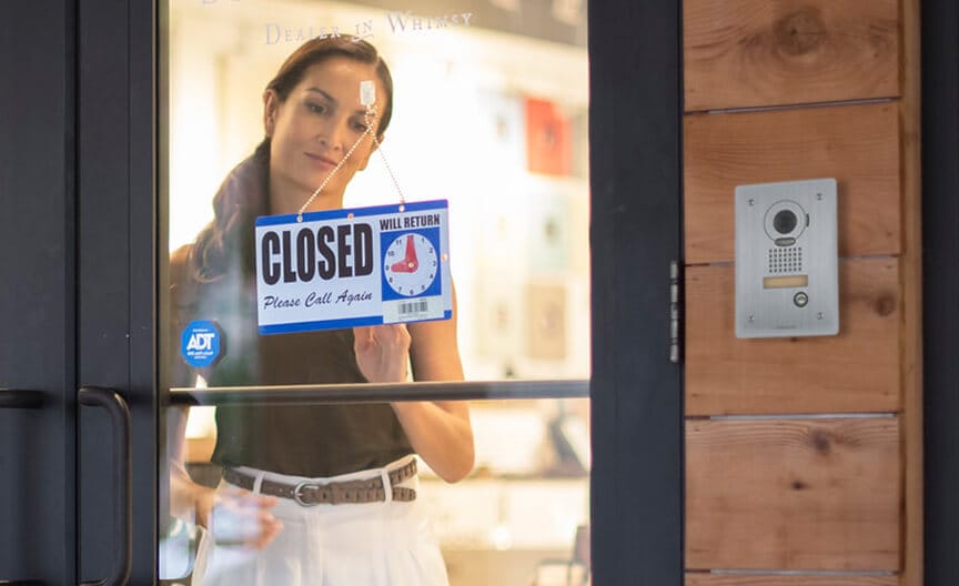 A business owner adjusts a “Closed” sign on a glass door, with an ADT sticker and intercom visible nearby.