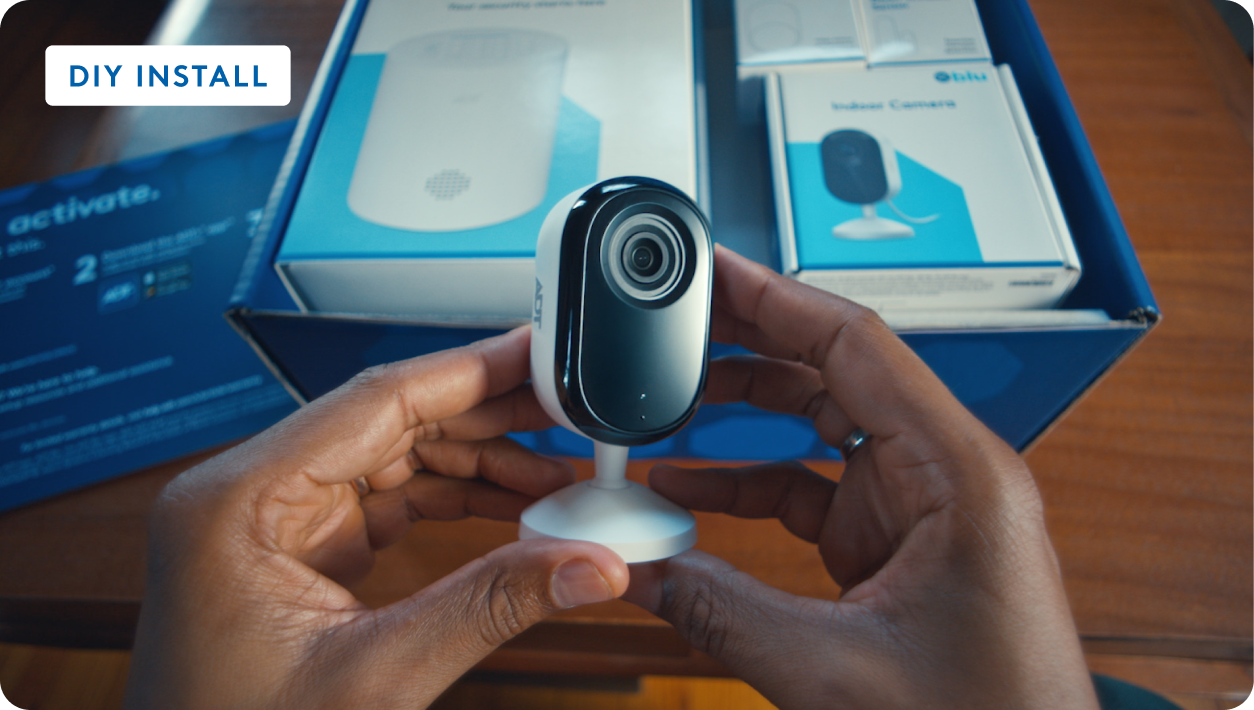 Hands holding a white indoor security camera over an open box of Blue by ADT components, labeled DIY Install.