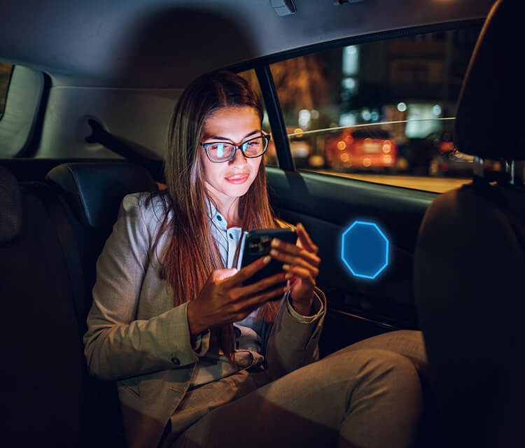 Traveling woman alone in the back of a rideshare at night looking at her smart phone. There is a glowing ADT emblem next to her, indicating the new My Safety service in the ADT+ app.