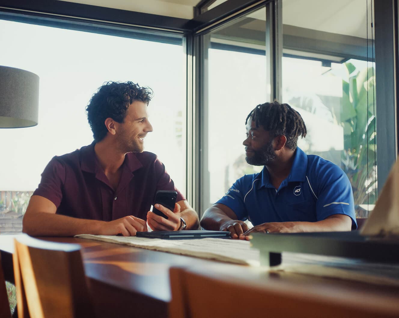 Paquete de ADT en un estante Un profesional de ADT se sienta en una mesa con un propietario, información profesional sobre la instalación en un dispositivo móvil.