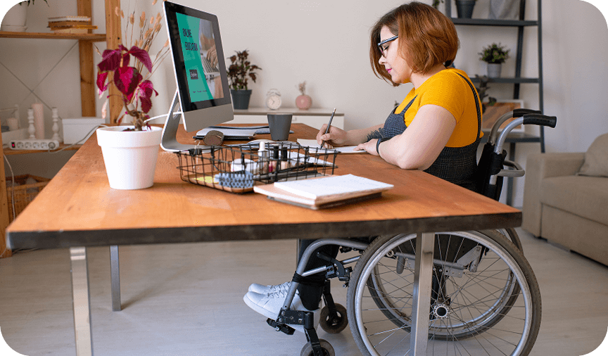 Woman using a wheelchair working at a desk, writing in a notebook beside a desktop computer.