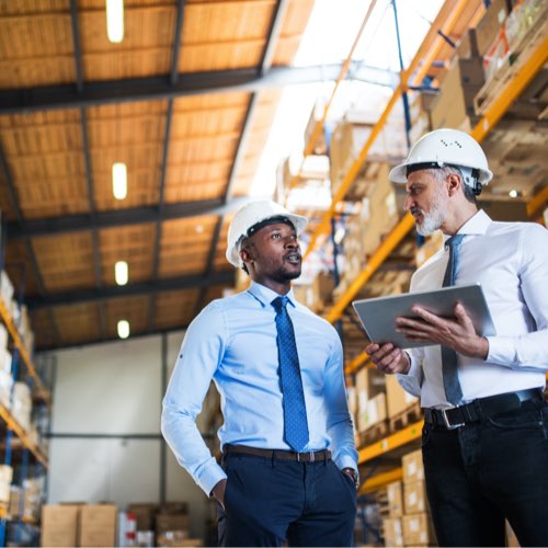 Two men with hardhats, one holding a tablet, in a warehouse