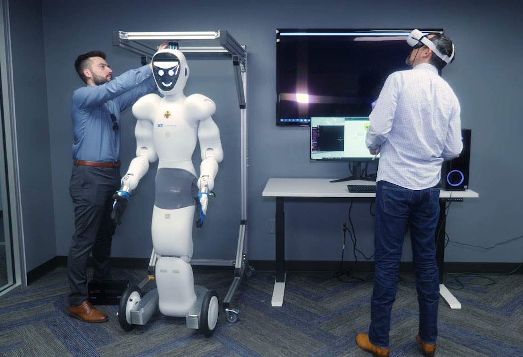 Man wearing VR goggles while another man makes adjustments to a robot