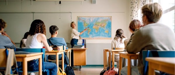 Students at desks looking at teacher pointing at world map