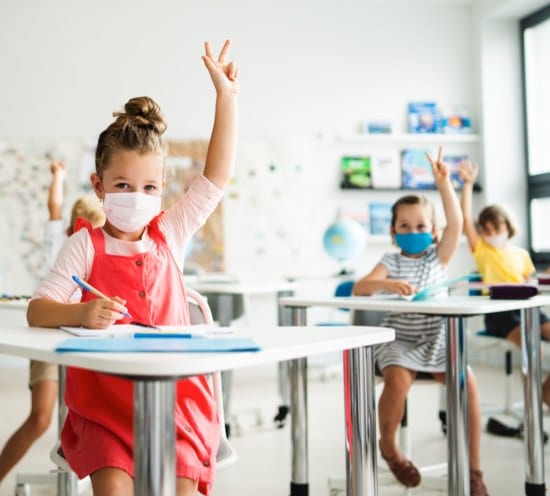 Children with raised hands wearing COVID-19 masks in a classroom