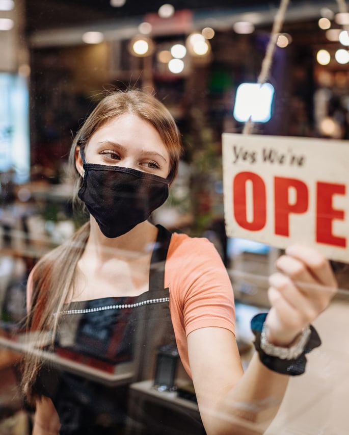 Store worker wearing a COVID mask and turning the store sign to read "Yes, We're Open"