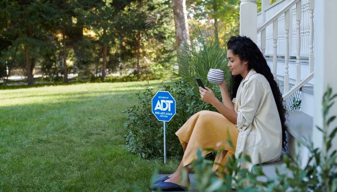 Woman sitting on her porch drinking coffee and looking at the ADT app on her phone